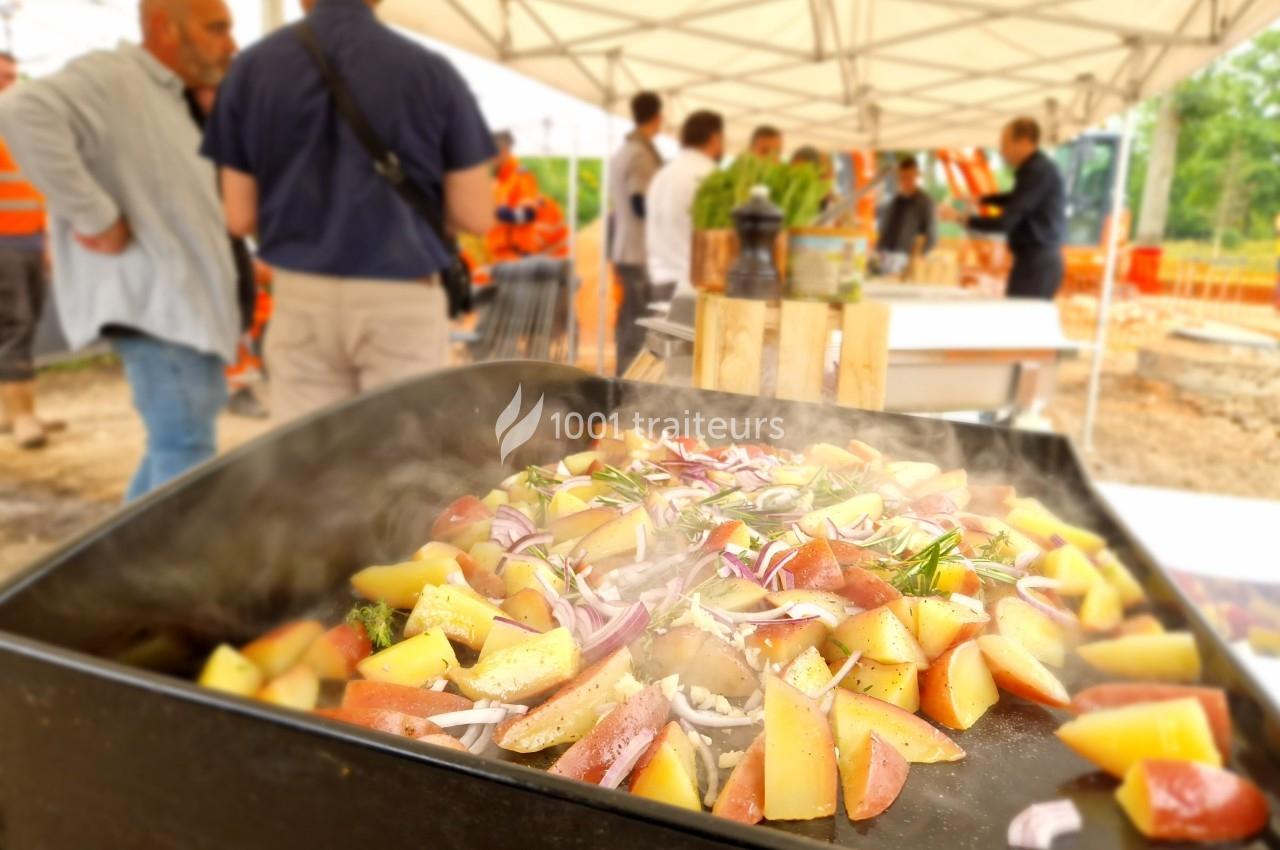 Pommes de terre et oignons rouges cuisinés sur une plancha fumante lors d'un événement en extérieur sous des tentes blanches.