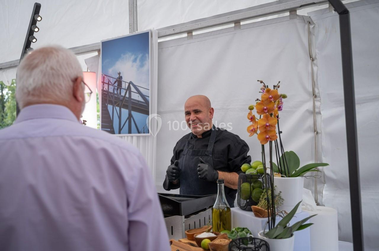 Un cuisinier souriant discute avec un homme devant un stand de cuisine décoré de fleurs et d'ingrédients frais.