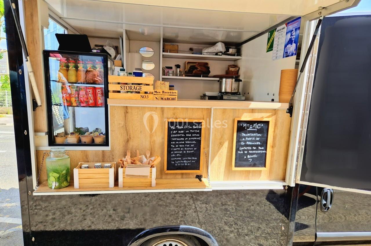 Camion de restauration avec comptoir en bois, ardoises affichant le menu, et étagères contenant boissons et accessoires.