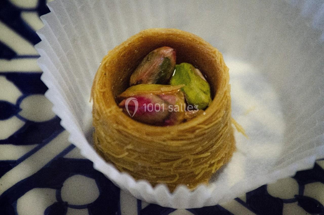 Pâtisserie orientale en nid de kadaïf garnie de pistaches, présentée dans une caissette en papier blanc.