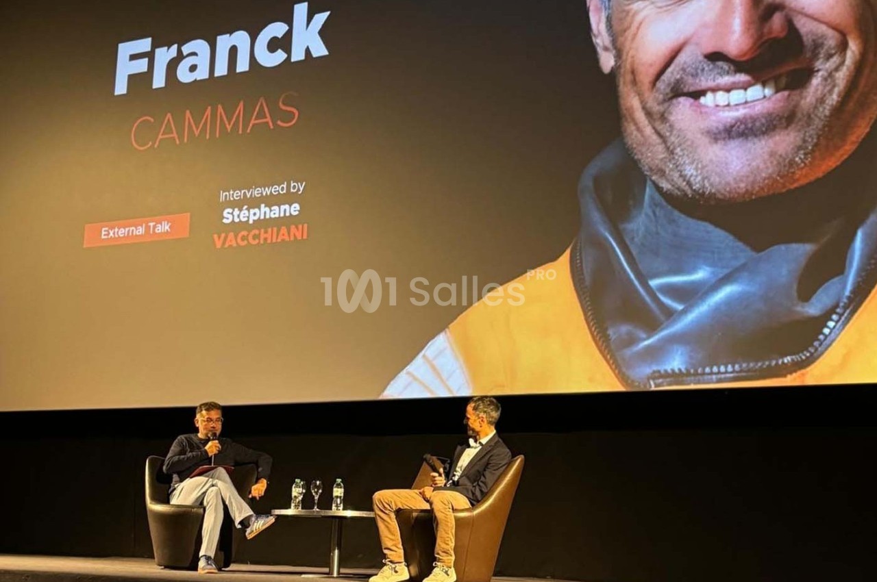 Deux hommes discutent sur une scène lors d'une interview, avec un écran affichant des informations sur Franck Cammas.