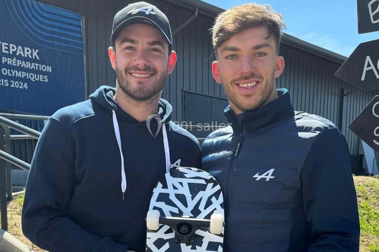 Photo Les Talents d'Espaces Réceptifs #4 Deux hommes souriants posent à l'extérieur, l'un tenant un skateboard noir et blanc devant un bâtiment moderne.