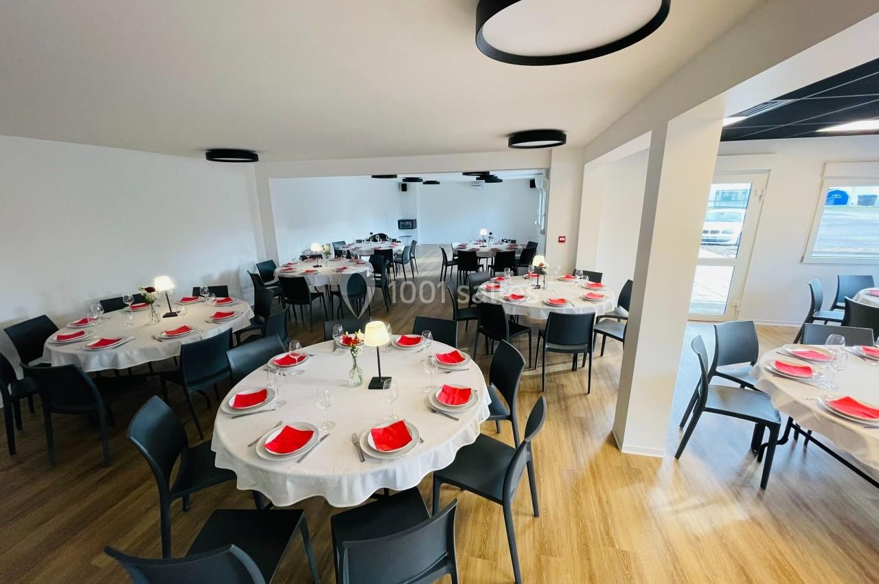 Salle de réception lumineuse avec tables rondes dressées de nappes blanches, serviettes rouges et chaises noires.