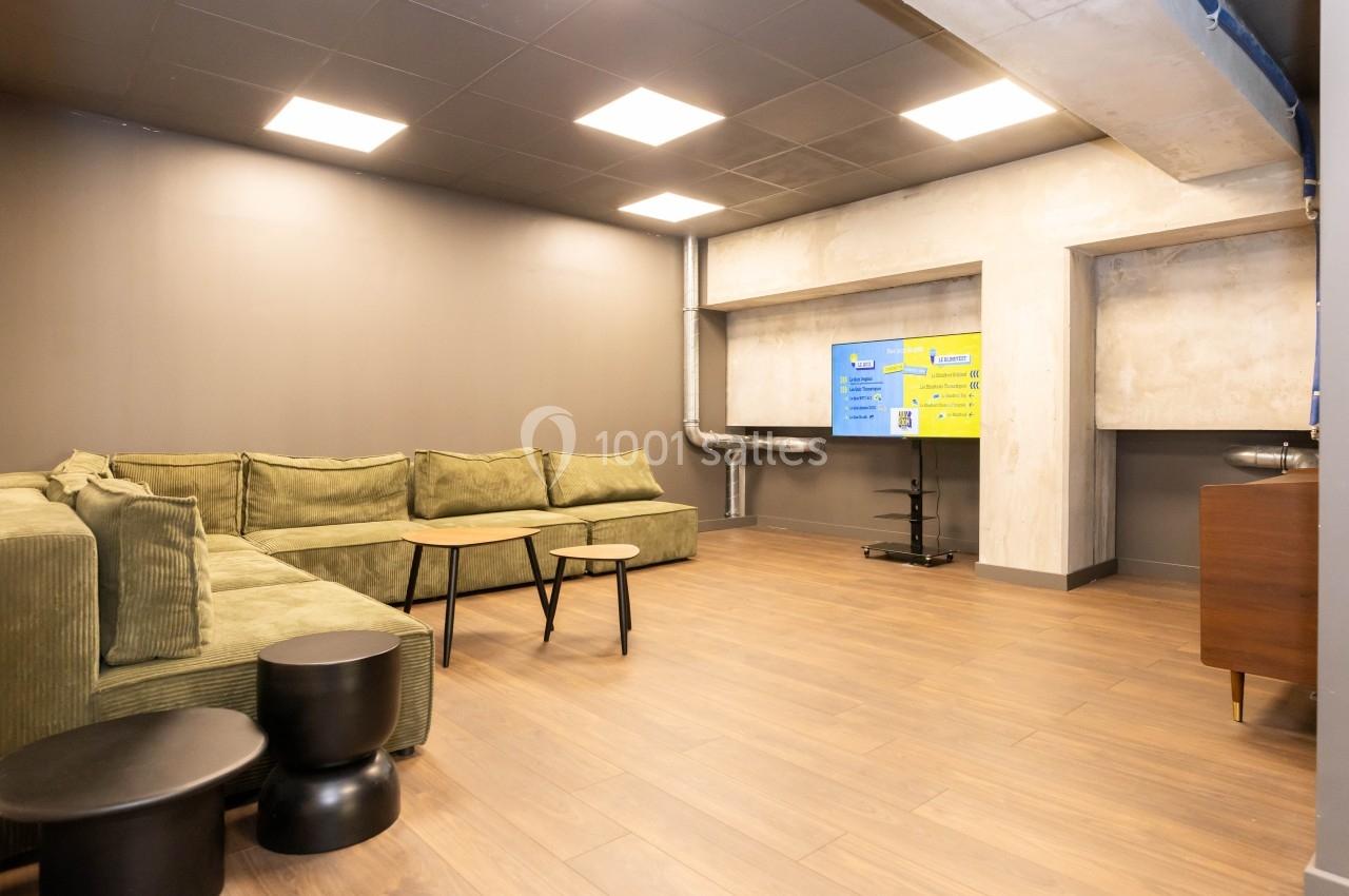 Salle lumineuse avec canapé vert, tables basses, écran mural et sol en parquet.