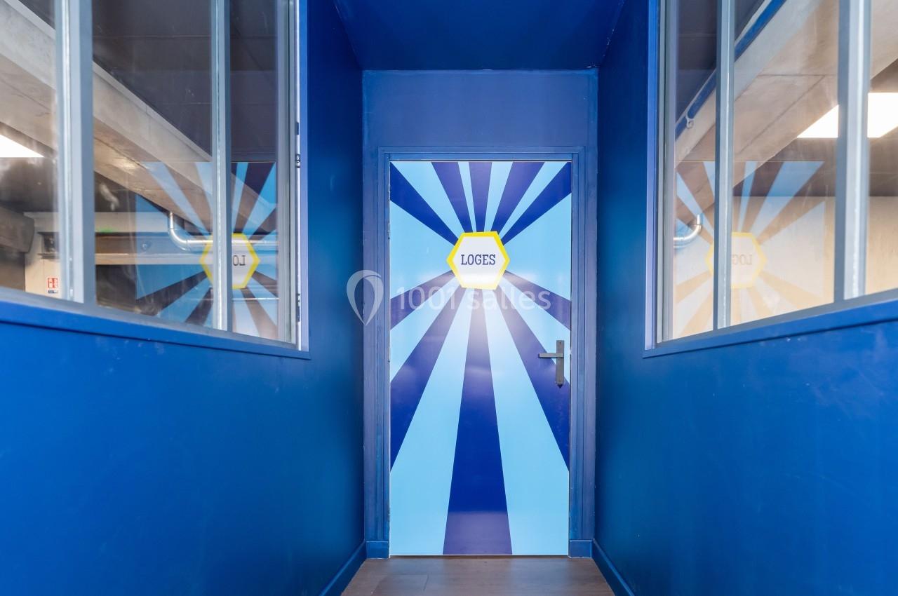 Porte bleue avec inscription ’Loges’, entourée de rayons lumineux peints, dans un couloir aux murs vitrés.