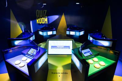 Poste de jeu interactif avec quatre boutons jaunes et un écran affichant ’Quiz Room’ dans un éclairage bleu.