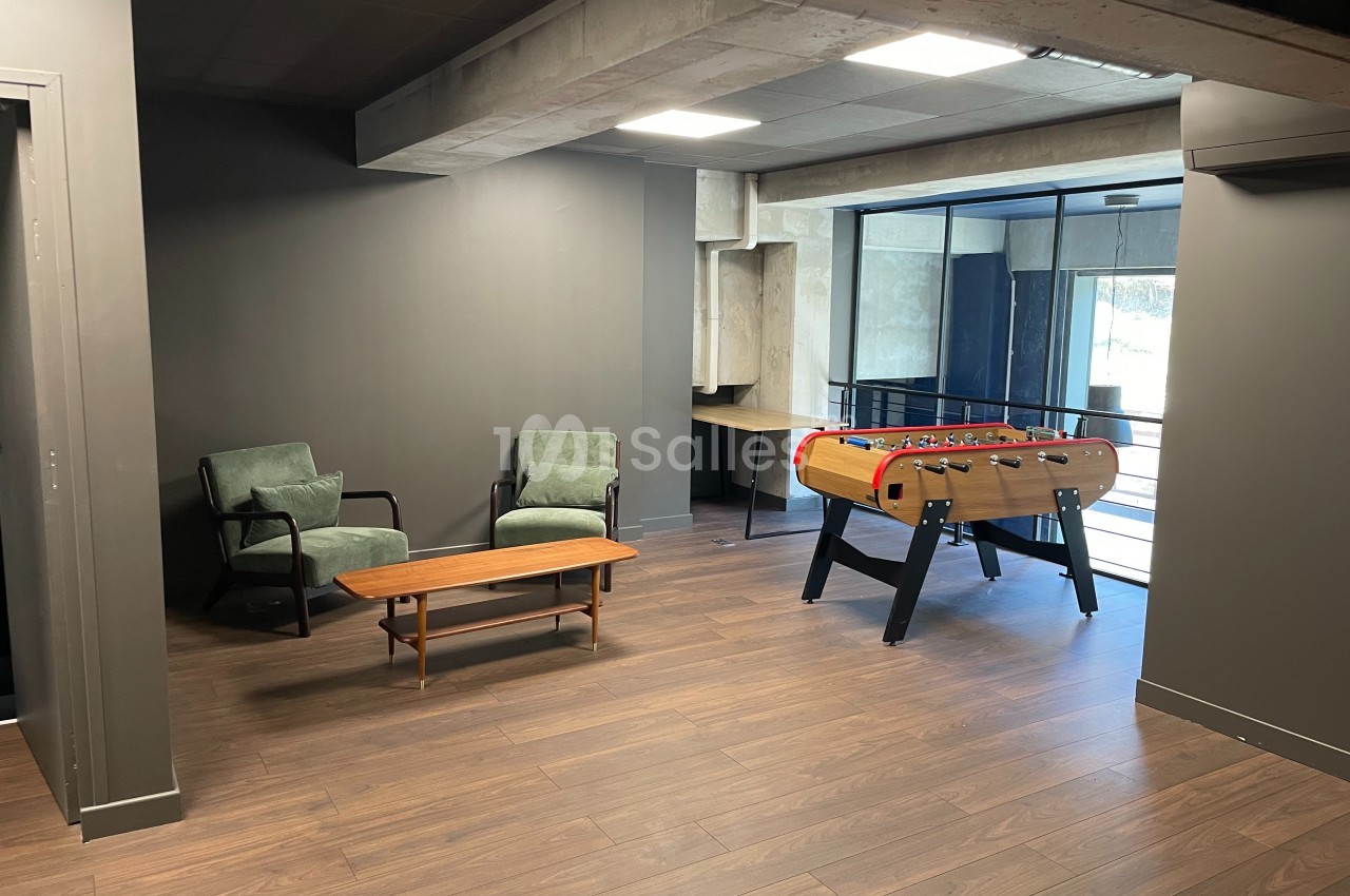Espace détente avec deux fauteuils, une table basse en bois et un baby-foot près d'une baie vitrée.