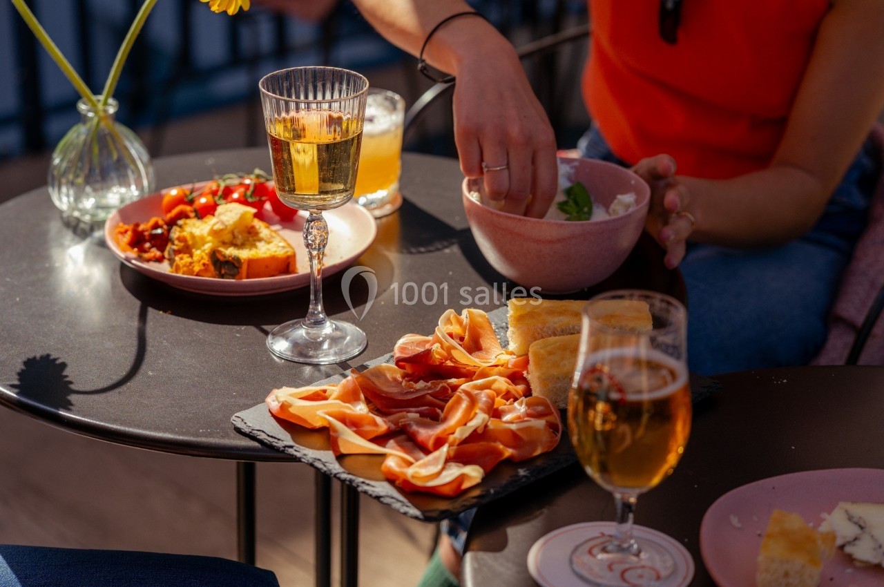 Assiette de charcuterie, pain et plats variés sur une table en extérieur, avec des boissons et une personne servant un bol.