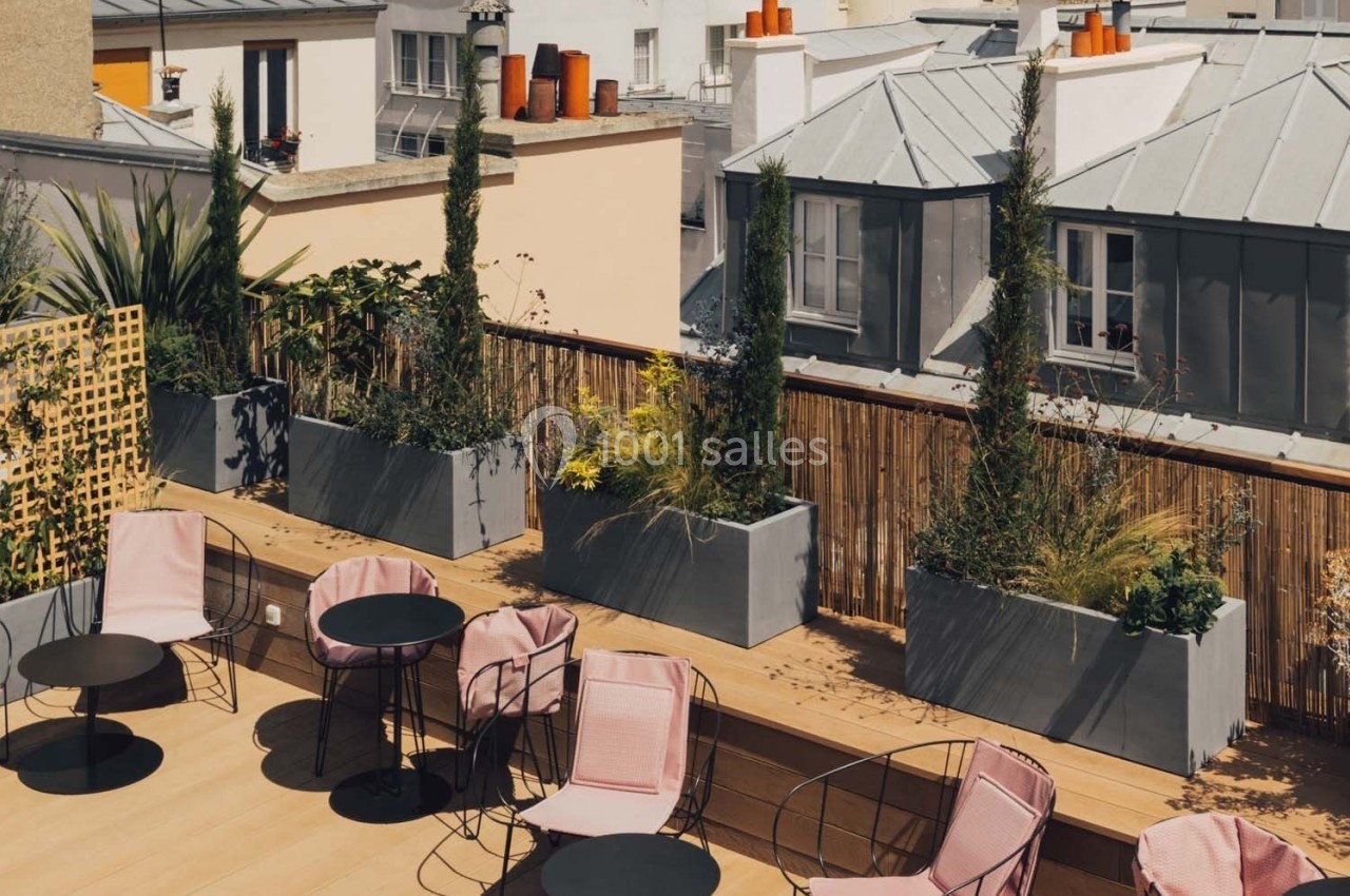 Terrasse ensoleillée avec des chaises roses, des tables noires et des jardinières devant des toits parisiens.