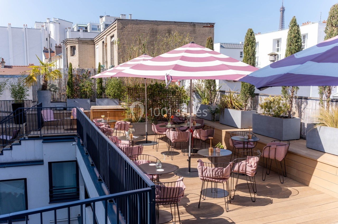 Terrasse ensoleillée avec tables, chaises et parasols rayés, entourée de plantes et de bâtiments urbains.