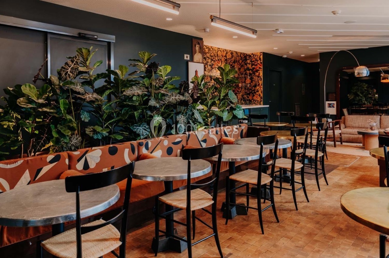 Salle de restaurant moderne avec tables rondes, chaises en bois et banquette décorée, entourée de plantes vertes.