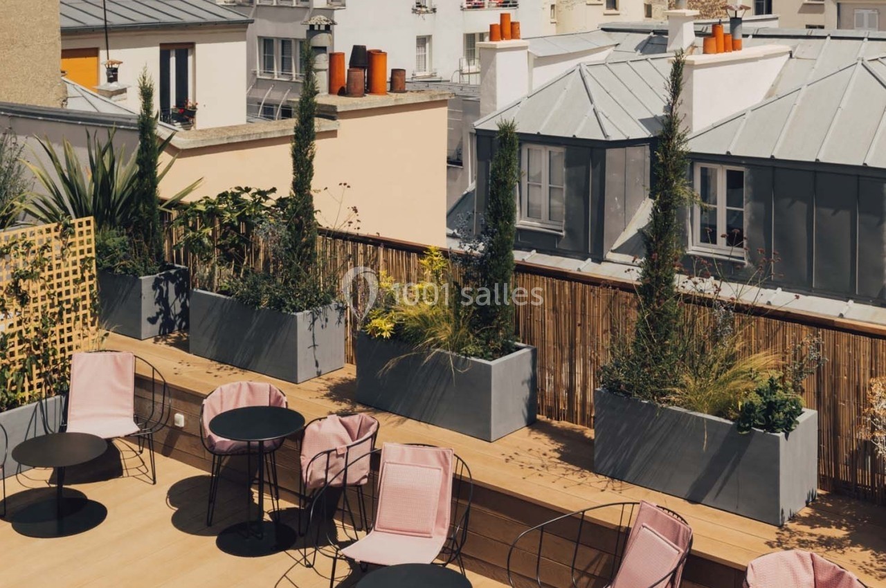Terrasse en bois avec tables, chaises roses et jardinières contenant des plantes, entourée de bâtiments urbains.
