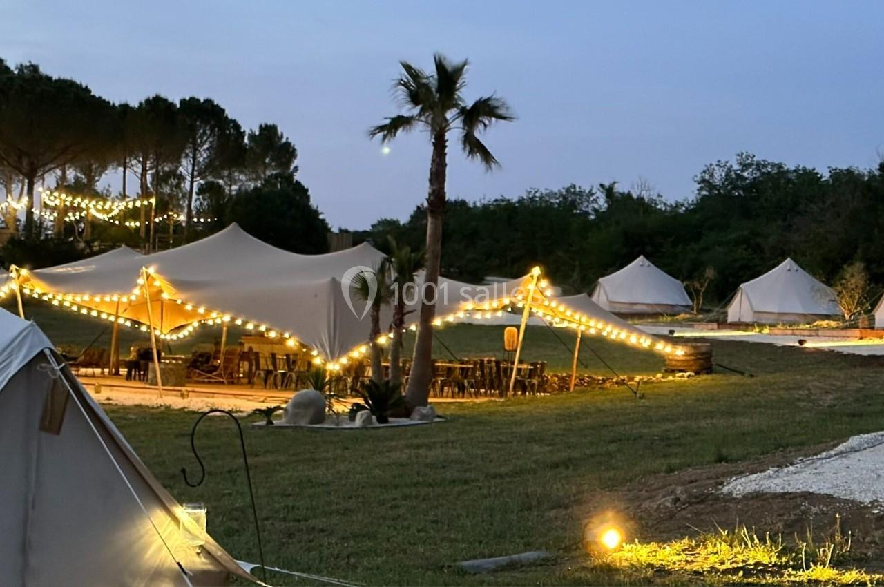Tentes éclairées par des guirlandes lumineuses dans un cadre naturel en soirée, avec arbres et pelouse.