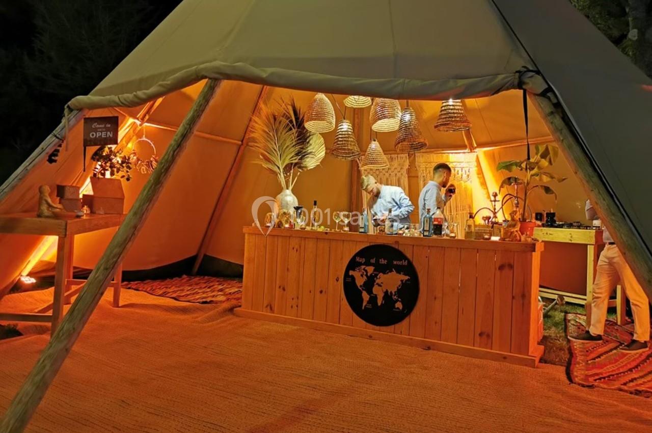 Intérieur d'une tente éclairée avec un bar en bois, des décorations suspendues et deux personnes préparant des boissons.