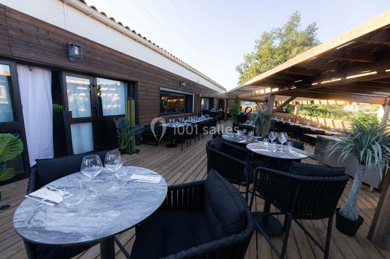 Terrasse en bois aménagée avec tables dressées, chaises noires et plantes, partiellement couverte par une pergola.