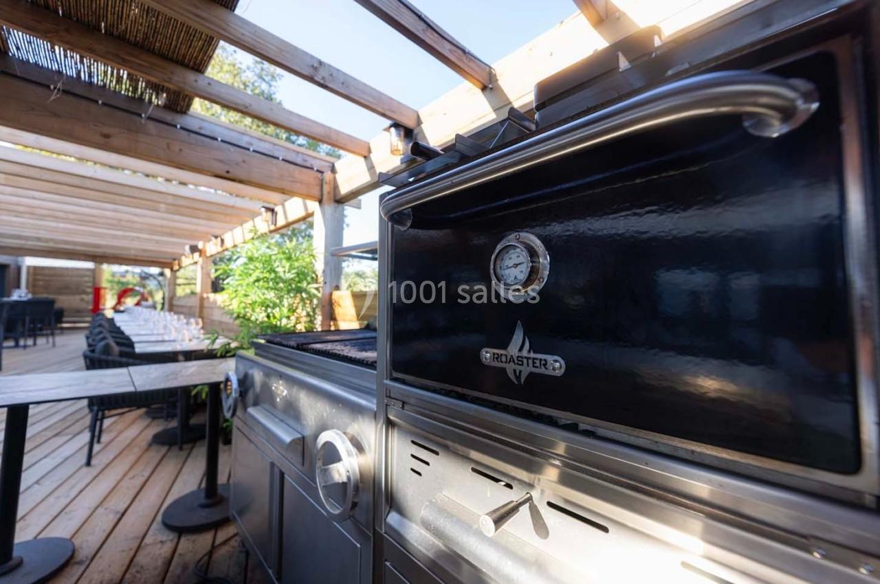 Barbecue noir sur une terrasse en bois, entouré de tables et d'une pergola avec lumière naturelle.