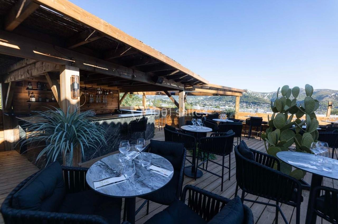 Terrasse en bois avec tables dressées, chaises noires, bar en arrière-plan et vue sur des collines ensoleillées.