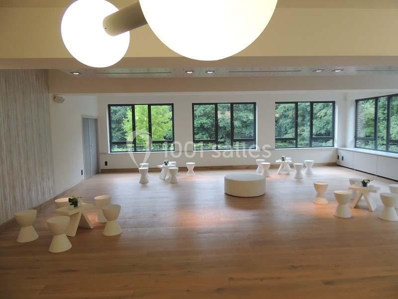 Salle lumineuse avec parquet, grandes fenêtres donnant sur des arbres, et mobilier blanc minimaliste disposé en cercle.