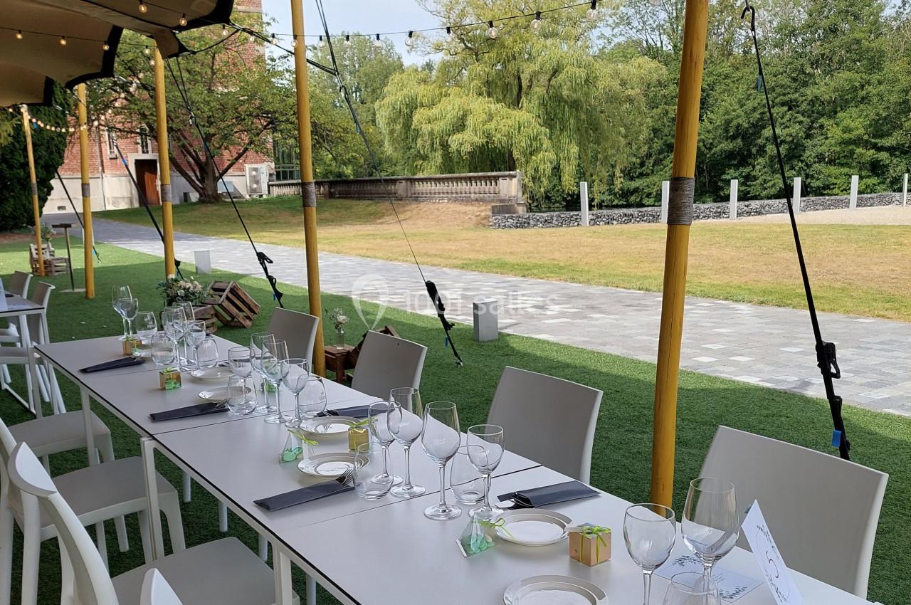 Table dressée en extérieur avec nappes blanches, verres et couverts, sous une tente avec vue sur un jardin verdoyant.