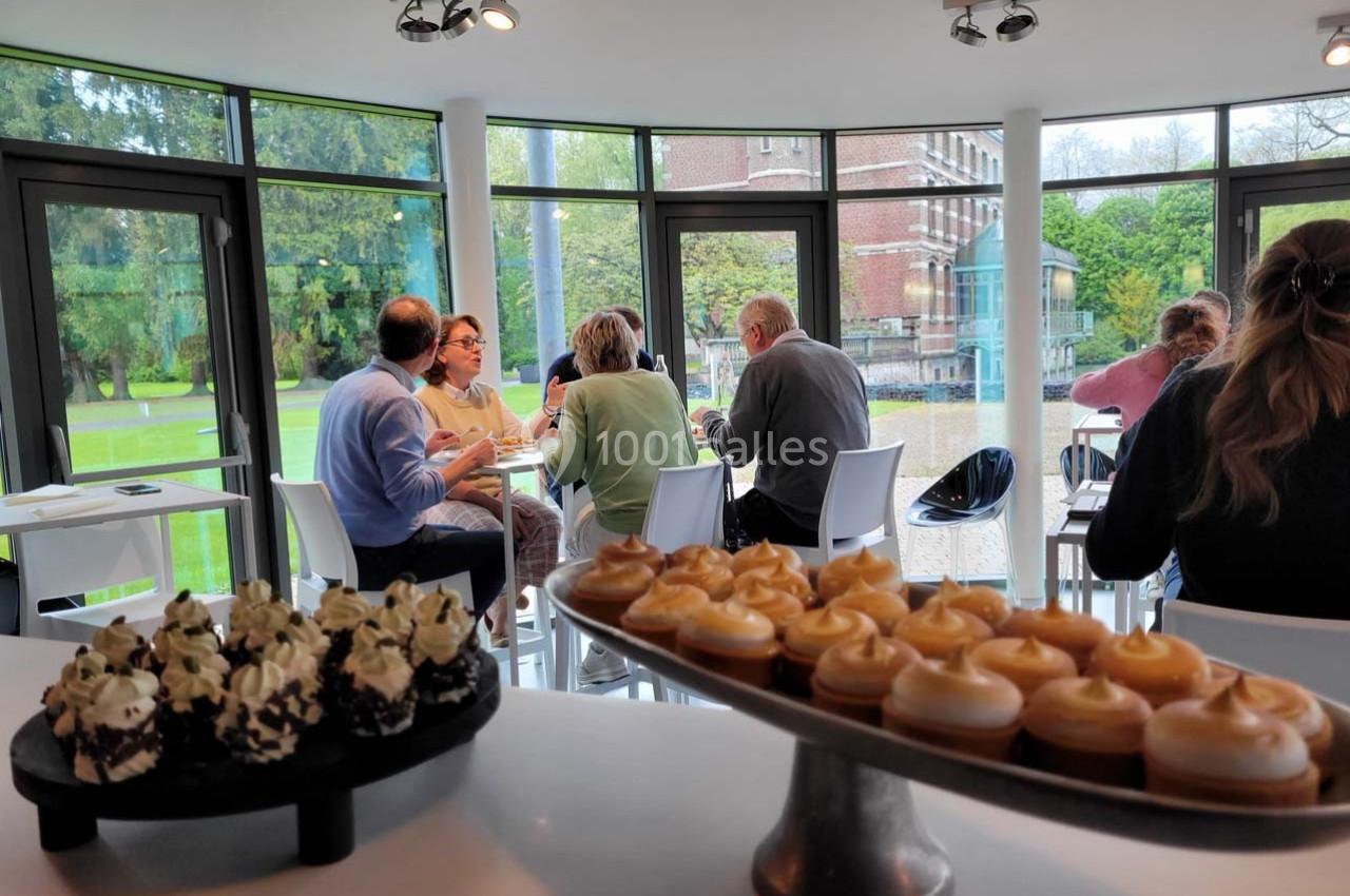 Des personnes discutent autour de tables dans une salle lumineuse avec vue sur un jardin, des desserts au premier plan.