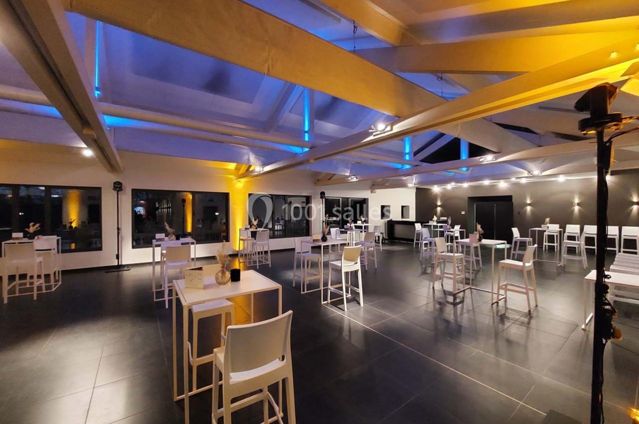 Salle spacieuse avec éclairage chaleureux, tables hautes blanches et grandes baies vitrées en arrière-plan.