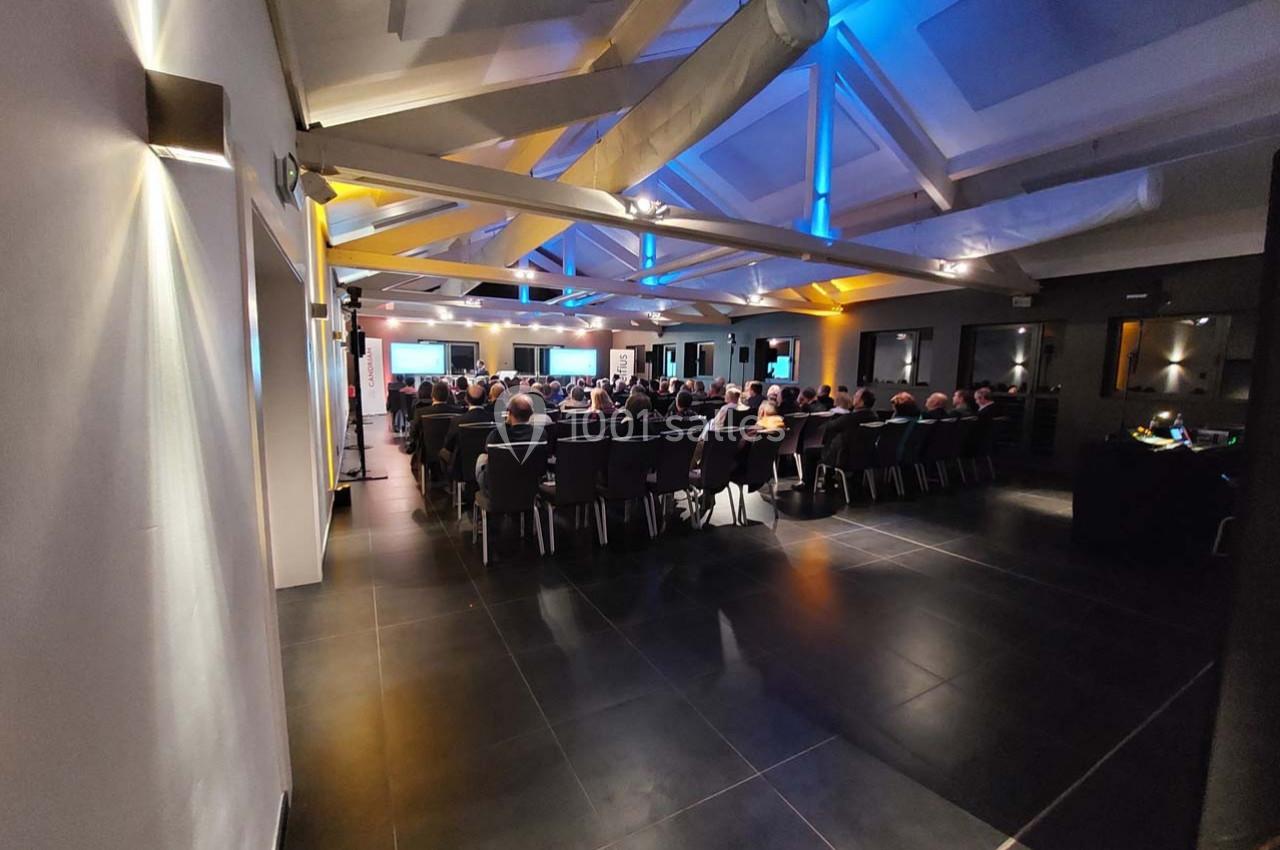 Salle de conférence moderne avec des participants assis, éclairée par des lumières bleues et jaunes au plafond.