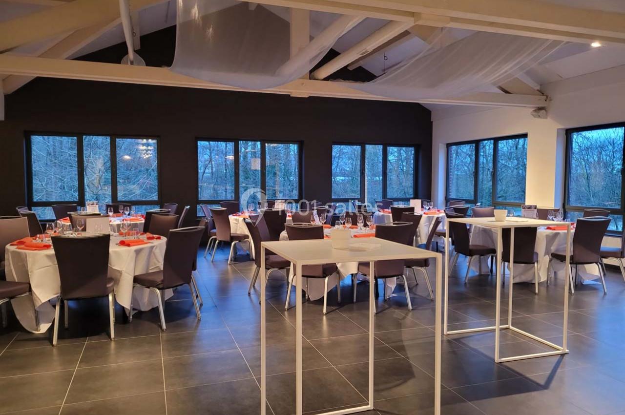 Salle de réception moderne avec tables rondes dressées, chaises violettes et grandes fenêtres donnant sur un paysage boisé.