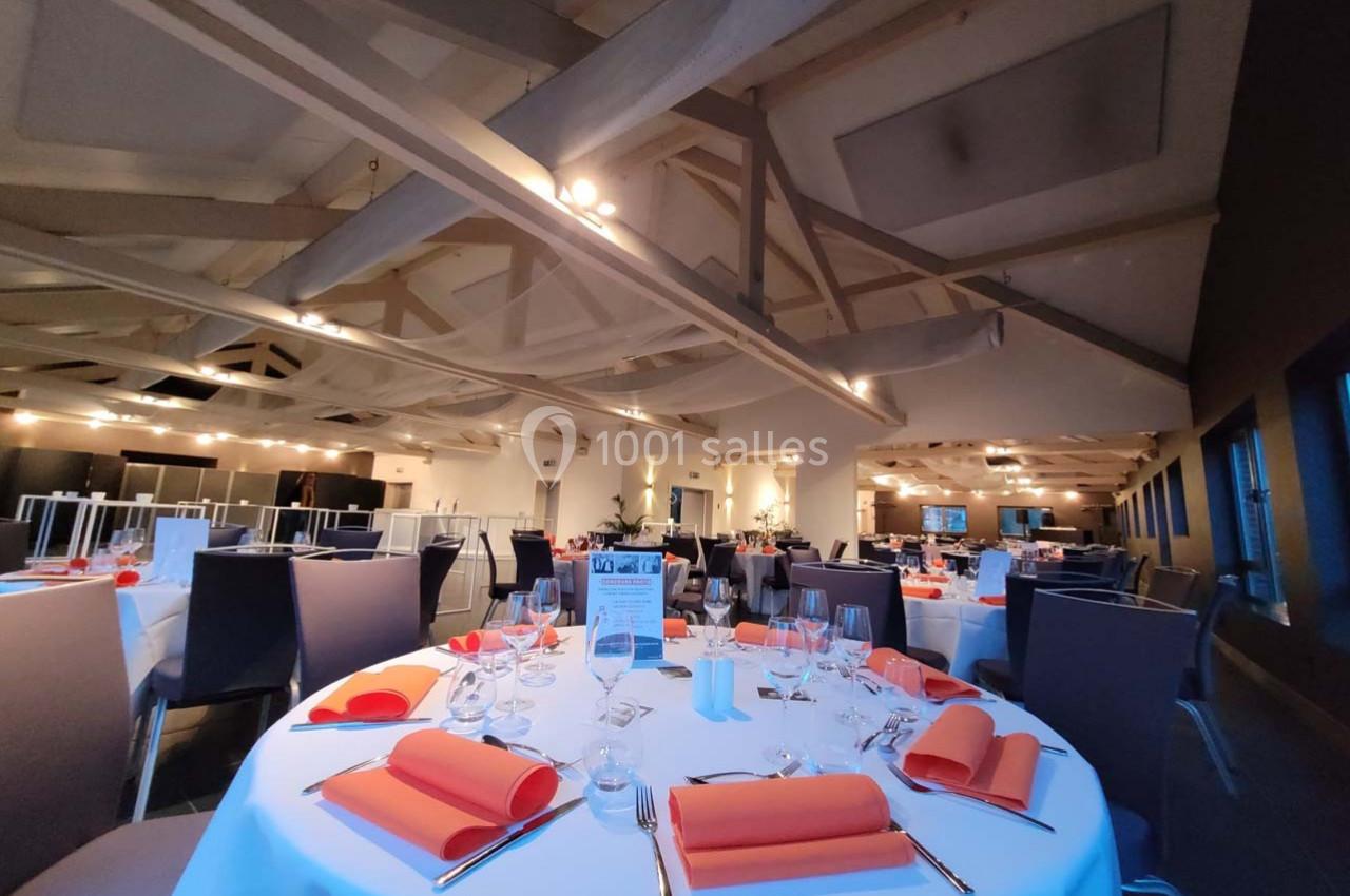 Salle de réception élégante avec tables rondes dressées, nappes blanches et serviettes orange, sous un plafond en poutres…
