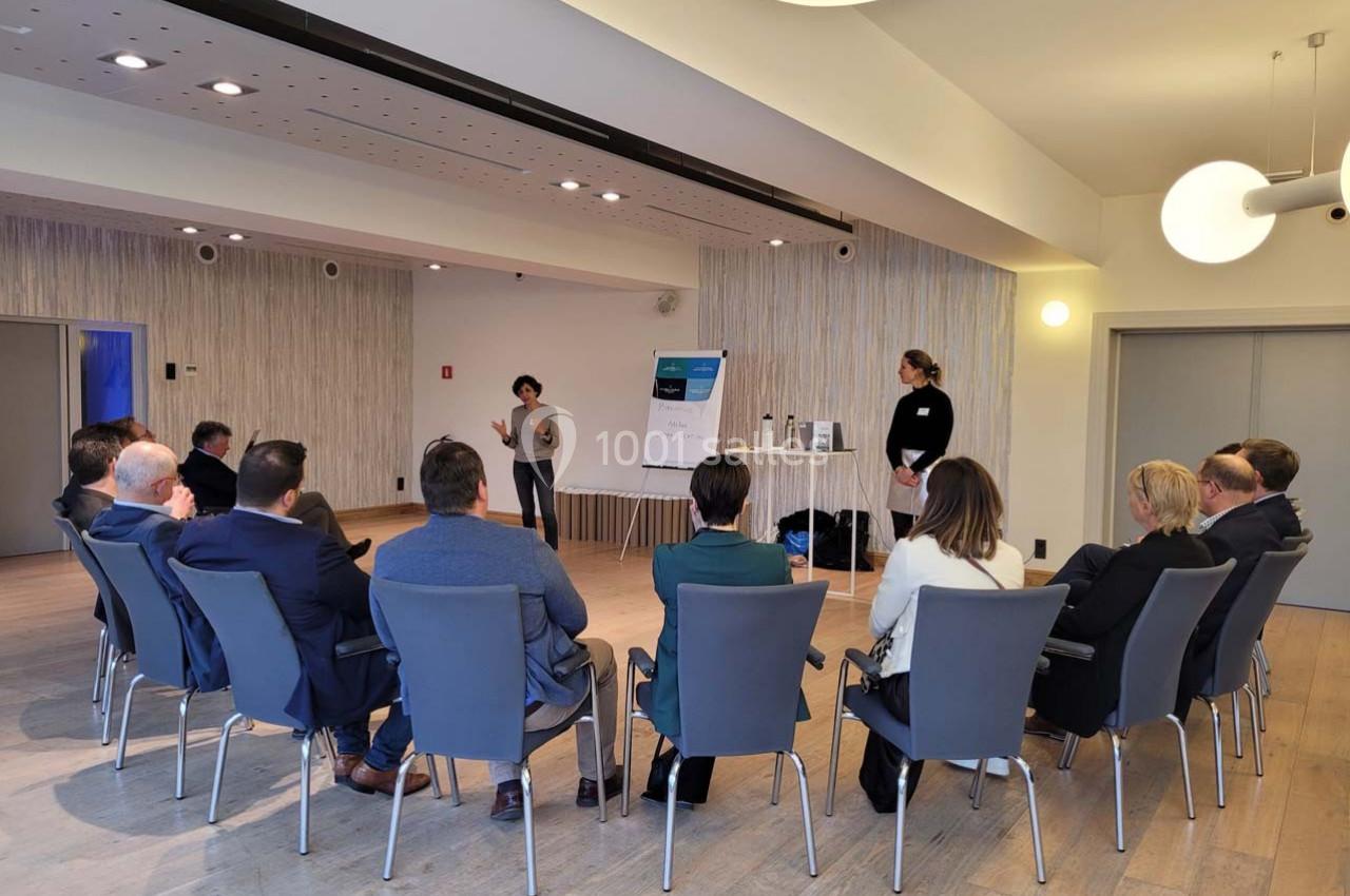 Une personne présente devant un groupe assis en cercle dans une salle lumineuse avec un tableau de conférence.