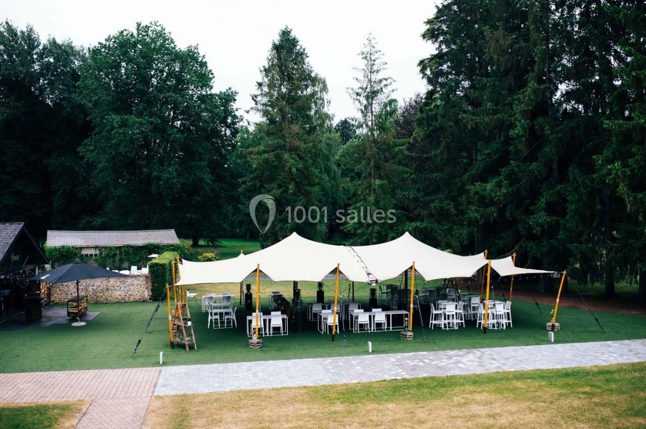 Tente blanche installée sur une pelouse entourée d'arbres, avec des tables et des chaises disposées en dessous.