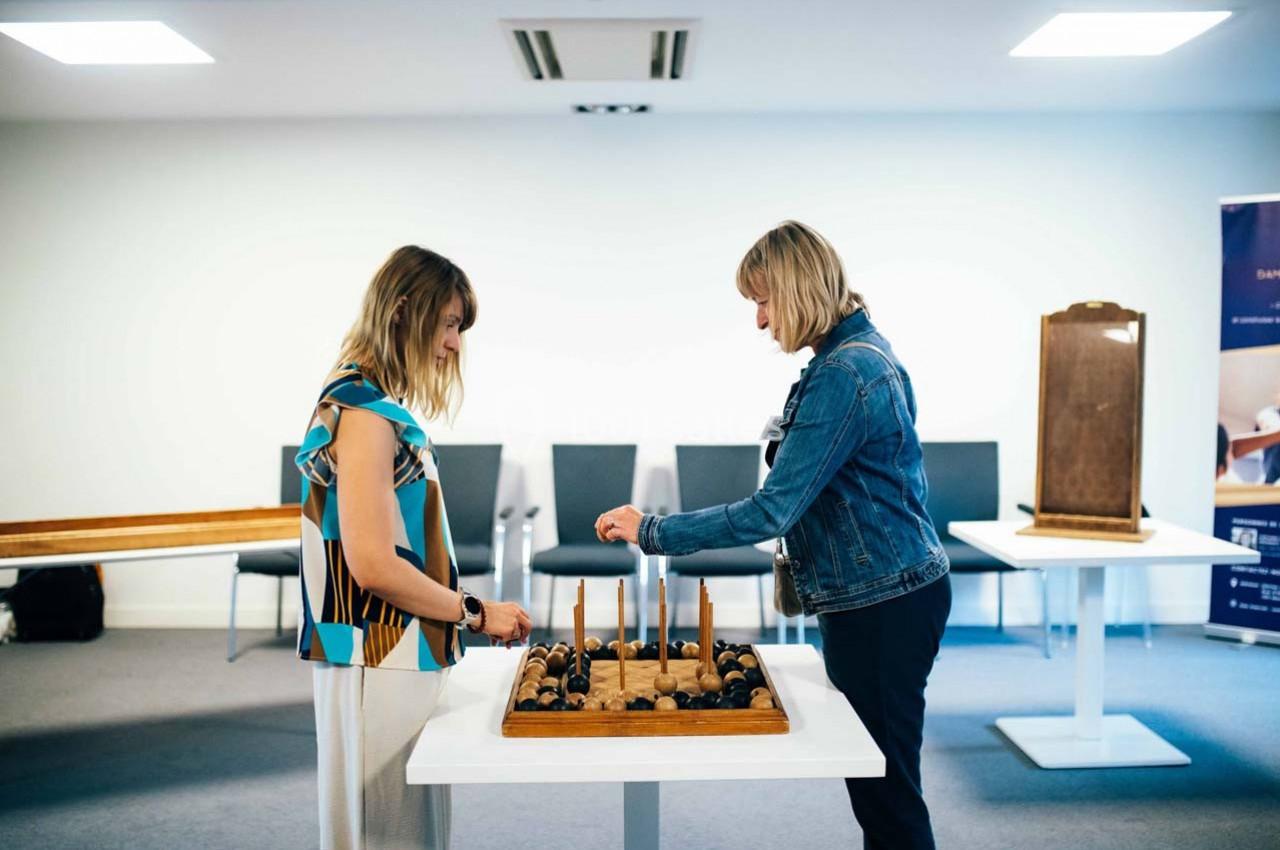 Deux femmes interagissent avec un jeu en bois sur une table dans une salle lumineuse avec des chaises en arrière-plan.