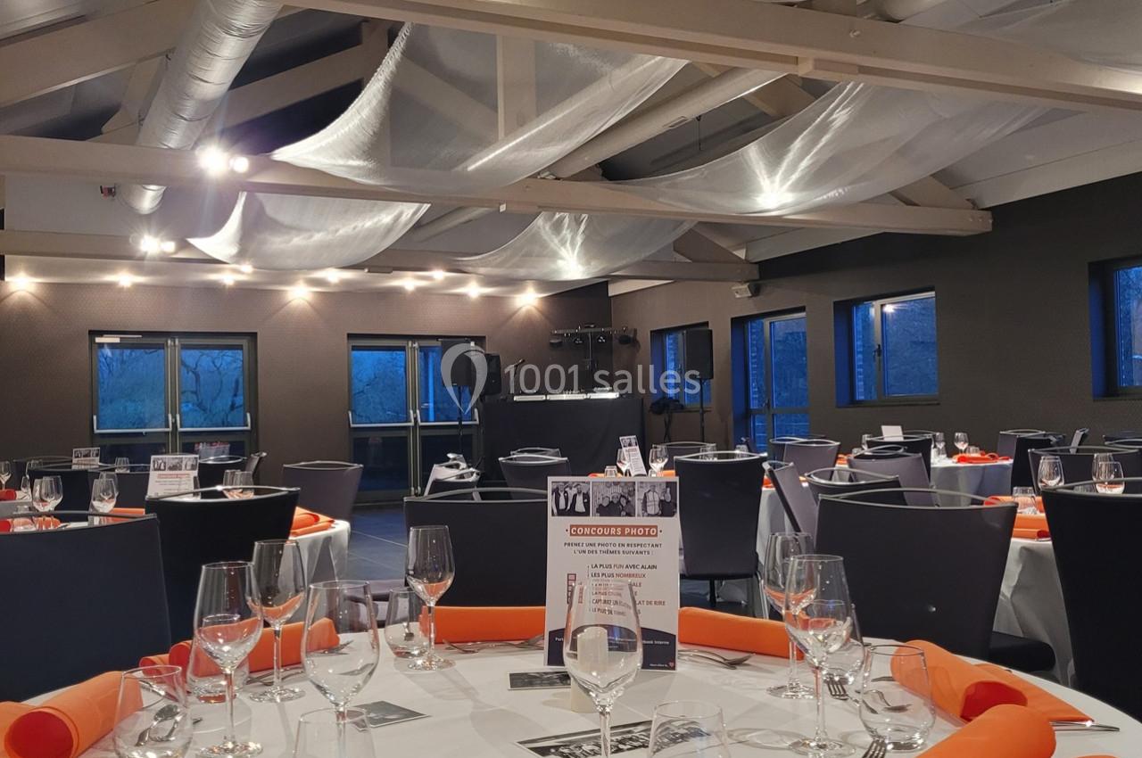 Salle de réception décorée avec des tables dressées, nappes blanches, serviettes orange et baies vitrées donnant sur l…