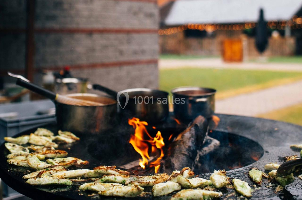 Légumes grillés sur une plaque chauffante au feu de bois, avec des casseroles en arrière-plan dans un espace extérieur.