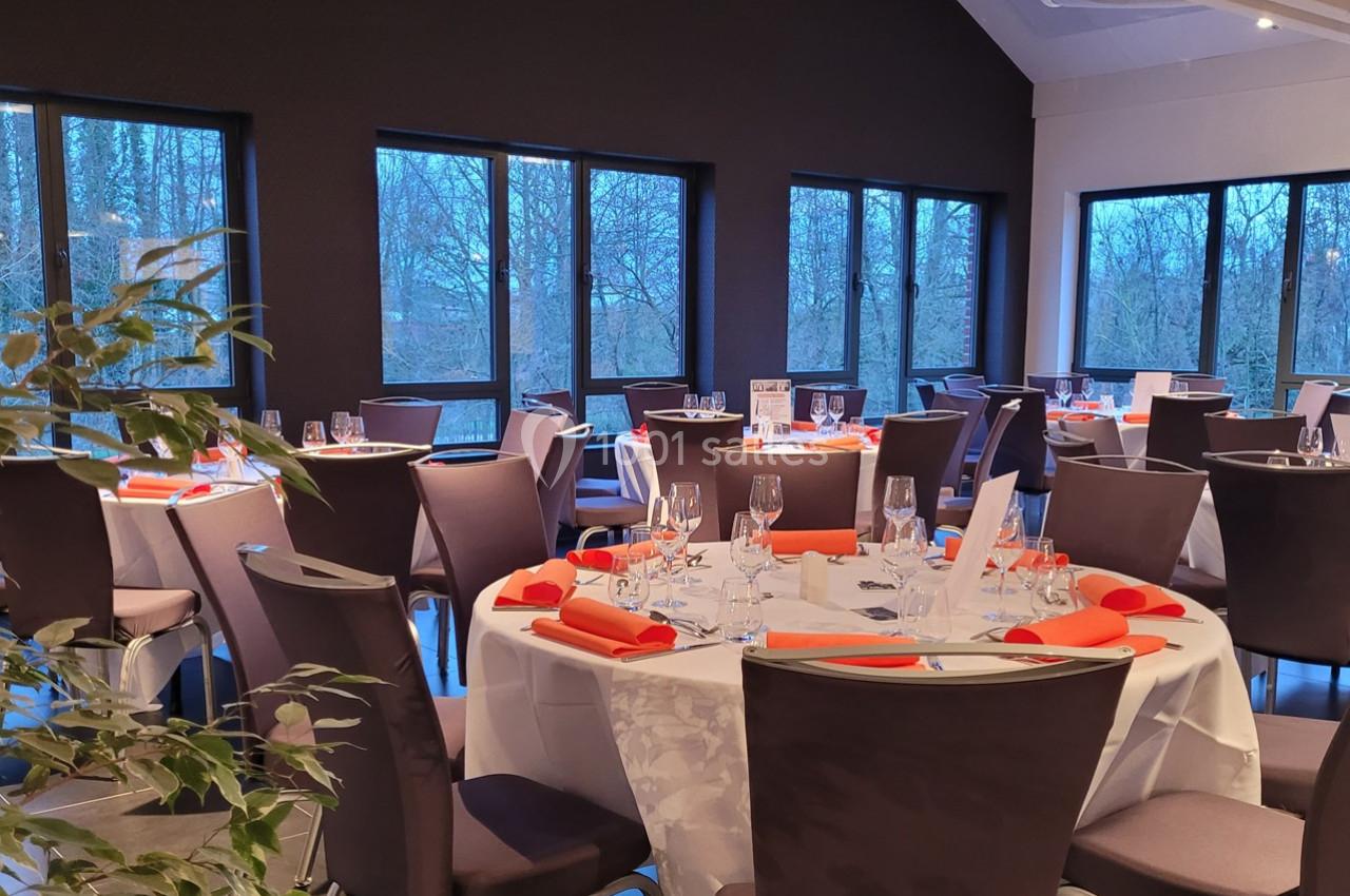 Salle de réception lumineuse avec tables rondes dressées, nappes blanches et serviettes orange, entourées de chaises grises.