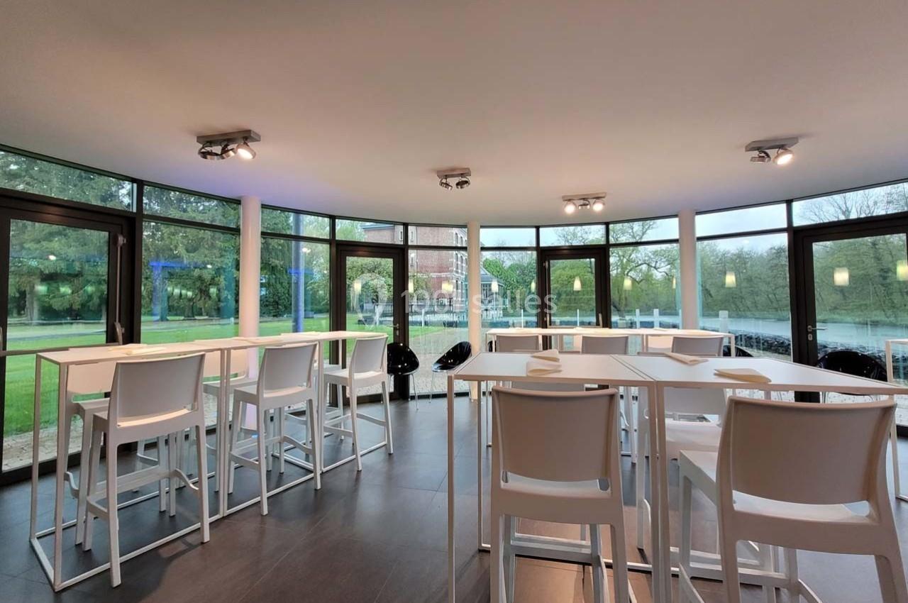 Salle lumineuse avec de grandes baies vitrées, tables hautes blanches et vue sur un jardin verdoyant.