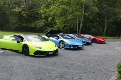 Quatre voitures sportives colorées alignées sur un parking en plein air, entourées de verdure.