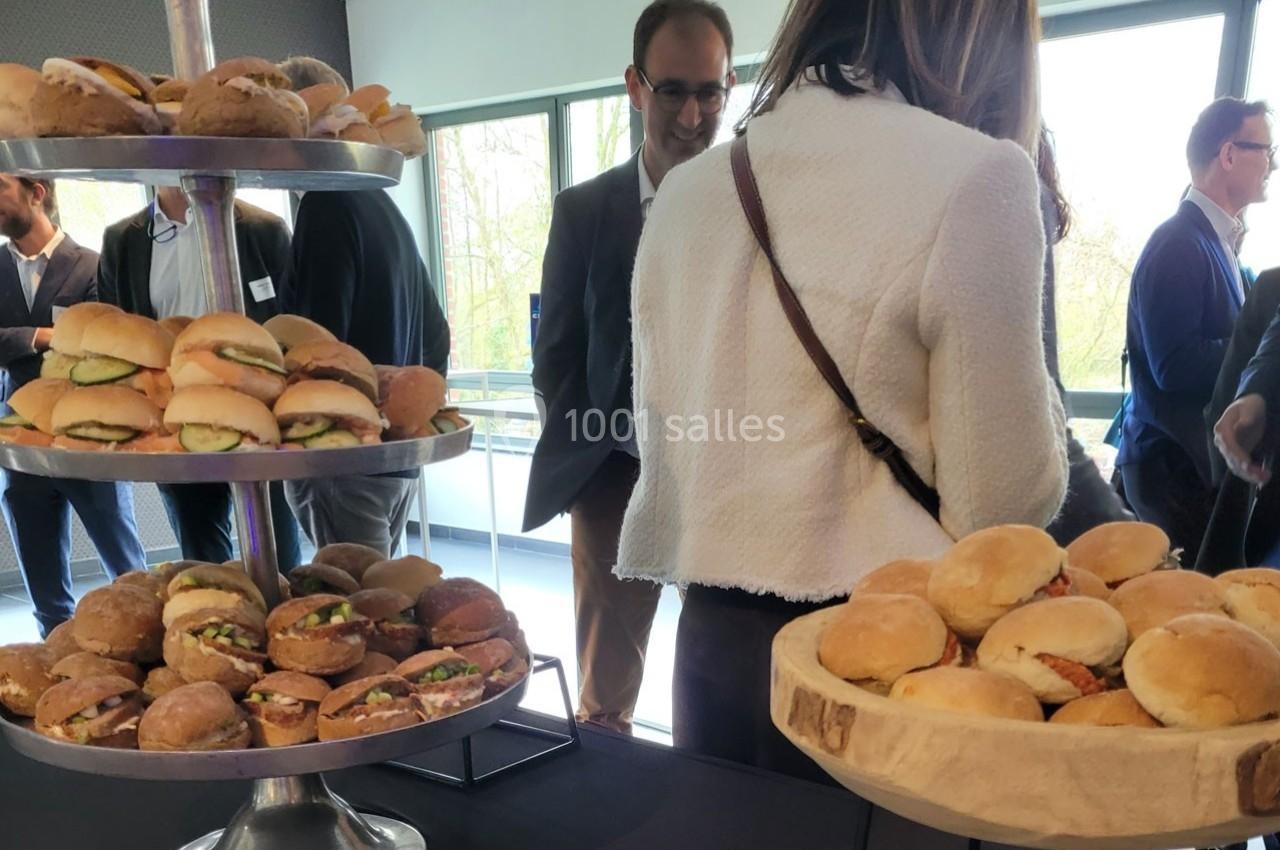 Plateaux de mini-sandwichs variés disposés sur une table lors d'un événement avec des participants en arrière-plan.