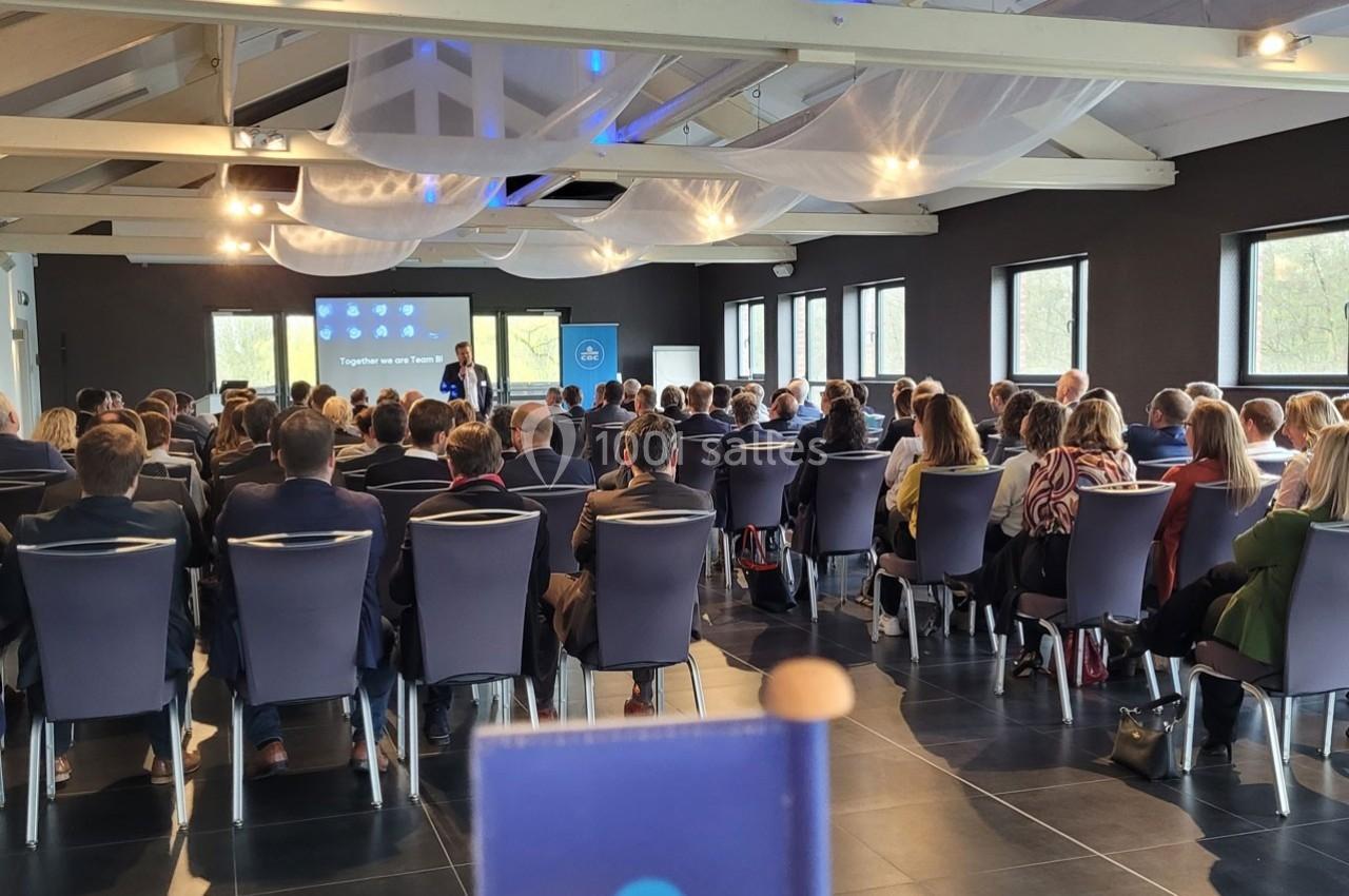 Un conférencier s'adresse à un public assis dans une salle lumineuse avec des écrans de présentation.