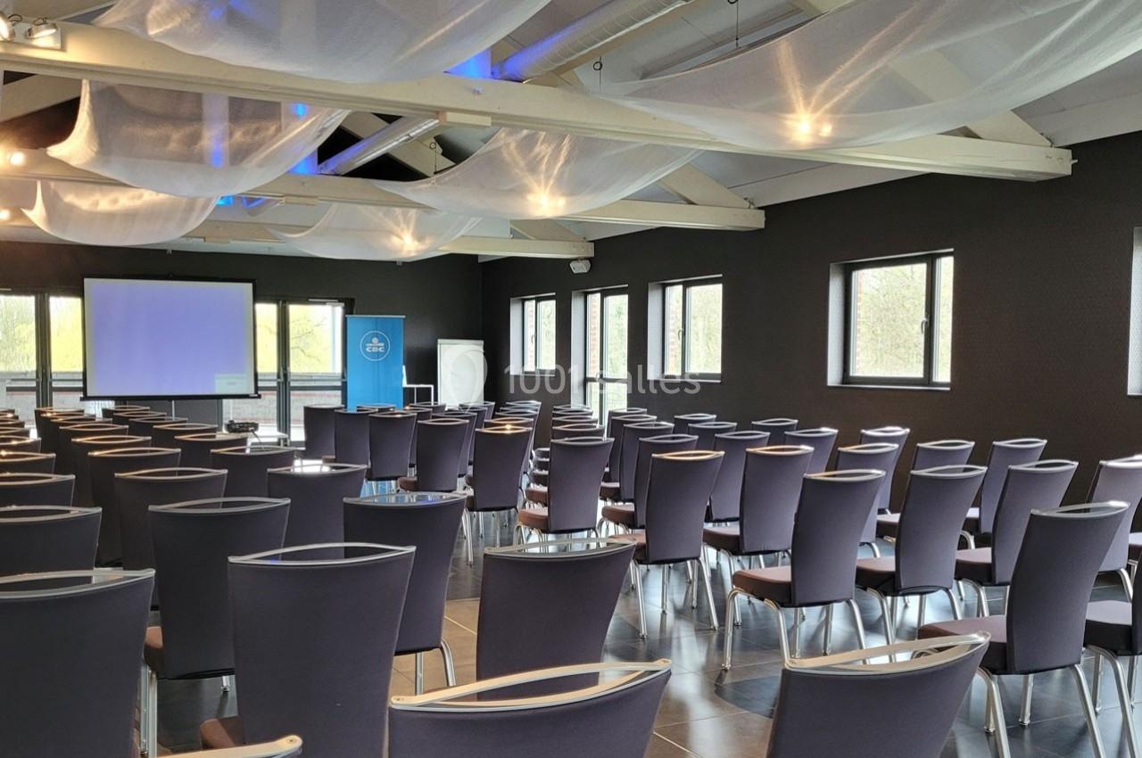 Salle de conférence lumineuse avec des rangées de chaises vides, un écran de projection et des fenêtres donnant sur l…