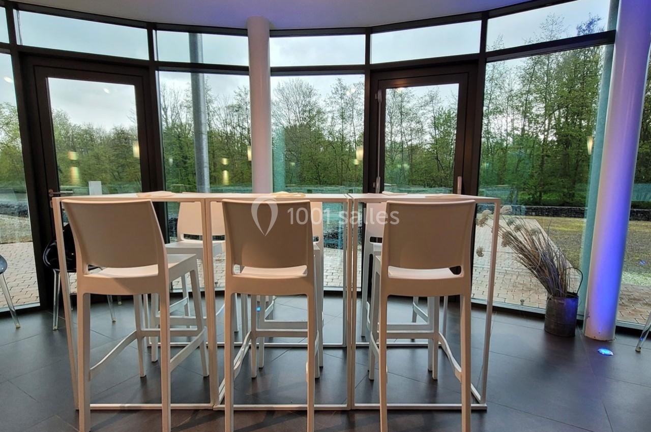 Quatre chaises hautes blanches autour de deux tables hautes, placées devant de grandes baies vitrées donnant sur un espace…