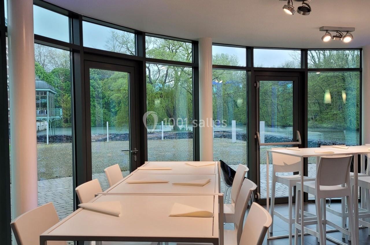 Salle lumineuse avec grandes baies vitrées donnant sur un espace extérieur arboré, tables et chaises blanches disposées.