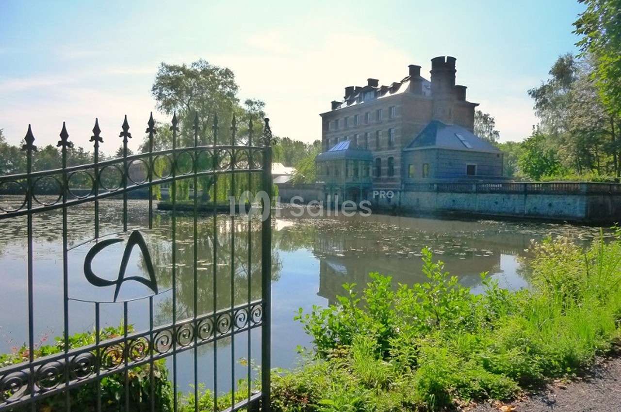 Manoir en pierre au bord d'un étang, entouré de végétation, avec une grille en fer forgé au premier plan.