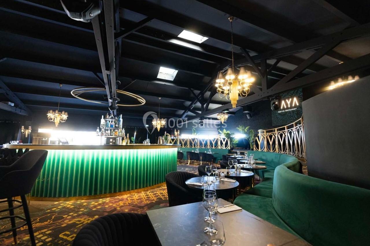 Intérieur d'un bar-restaurant moderne avec comptoir éclairé, fauteuils verts et lustres suspendus.