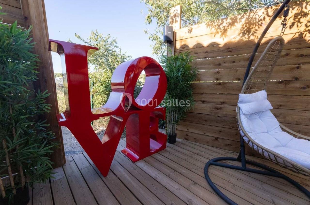 Grande sculpture rouge formant le mot ’LOVE’ sur une terrasse en bois entourée de plantes et d'un fauteuil suspendu.