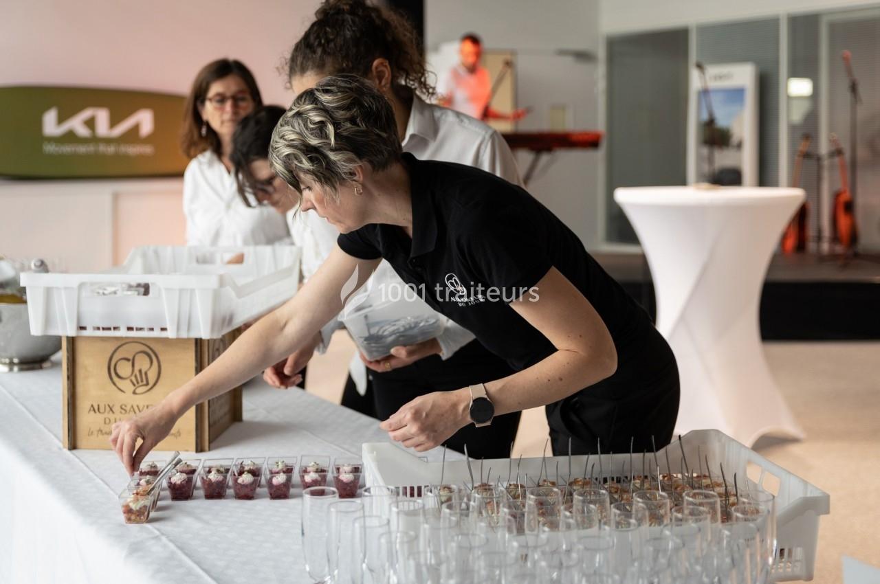 Une femme dispose des verrines sur une table lors d'un événement, avec d'autres personnes en arrière-plan.