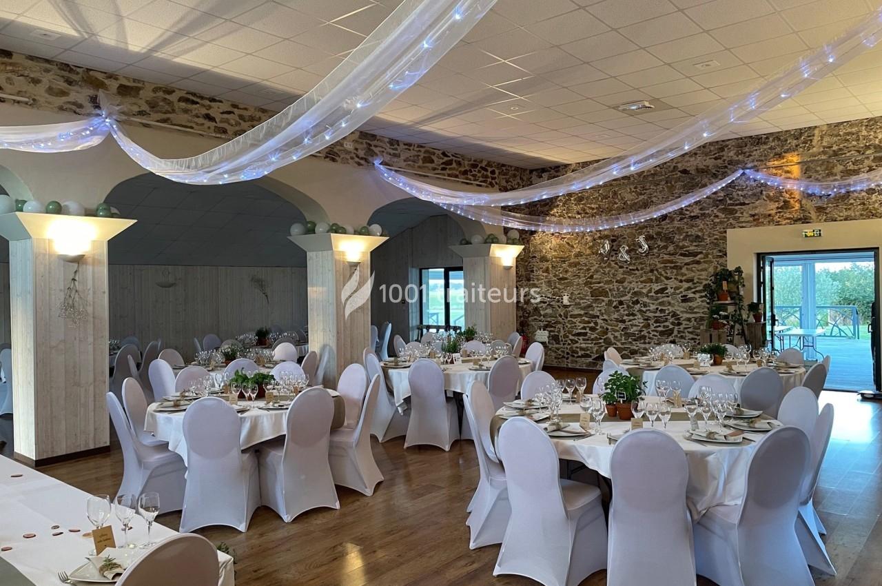 Salle de réception décorée avec des tables rondes dressées, des chaises blanches et des guirlandes lumineuses suspendues.