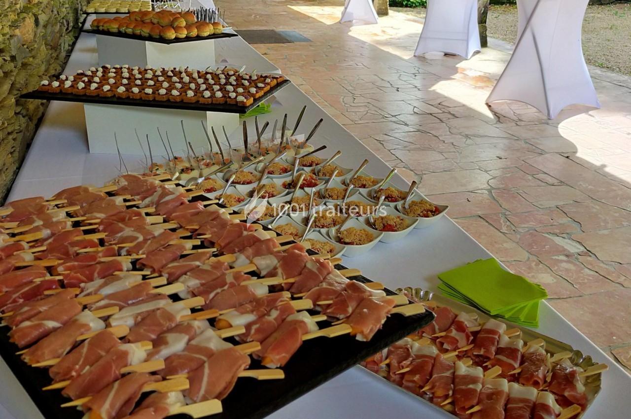 Buffet avec brochettes de jambon cru, verrines colorées et amuse-bouches disposés sur des tables en extérieur.