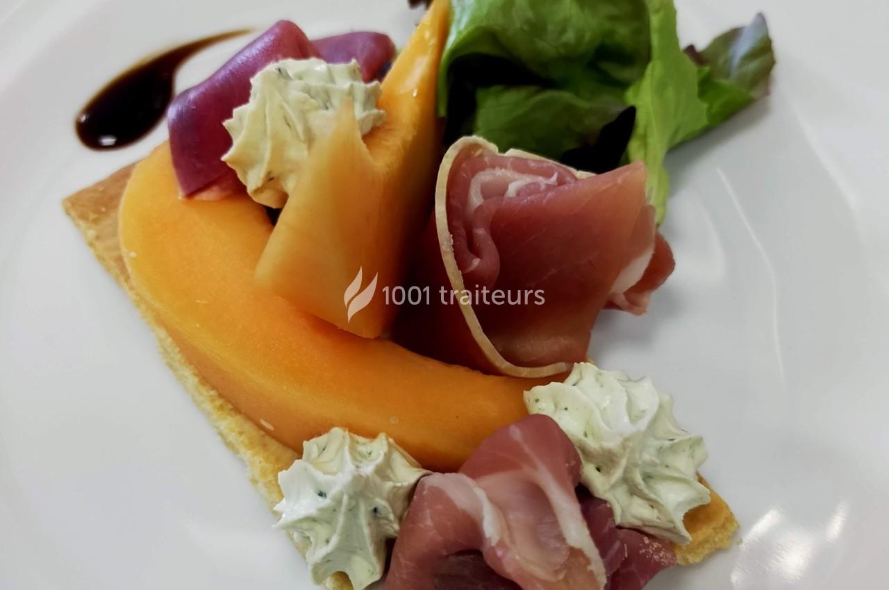 Tranche de melon avec jambon cru, crème aux herbes et salade verte sur une assiette blanche.