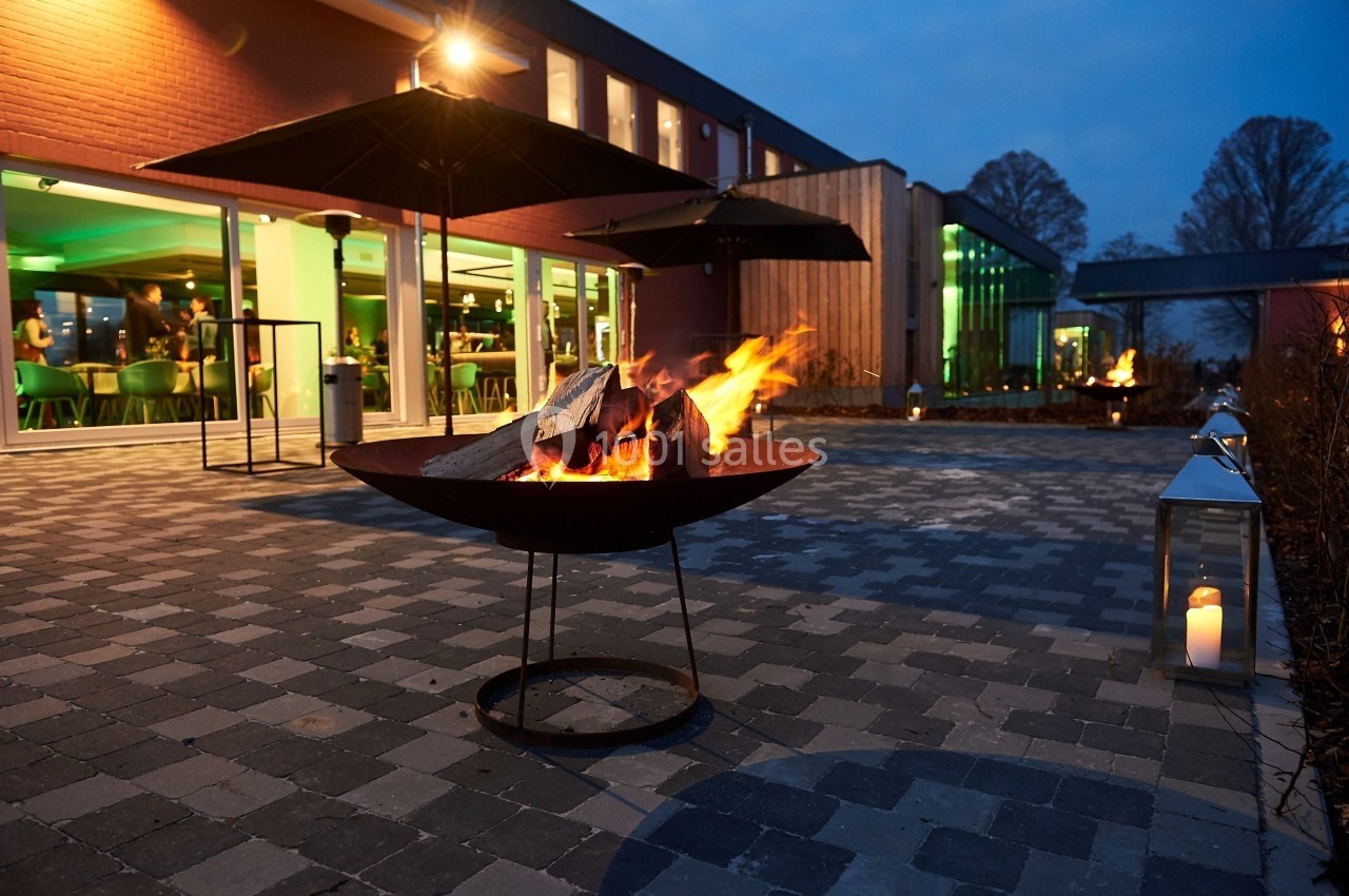 Brasero allumé sur une terrasse pavée en soirée, avec des bâtiments éclairés et des arbres en arrière-plan.