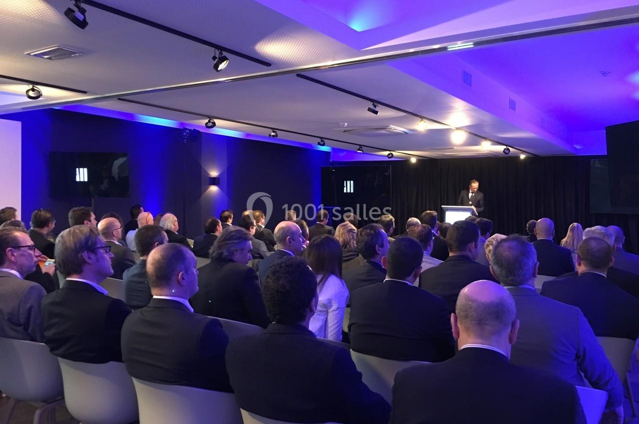 Un conférencier s'adresse à un public assis dans une salle moderne éclairée par des lumières bleues.